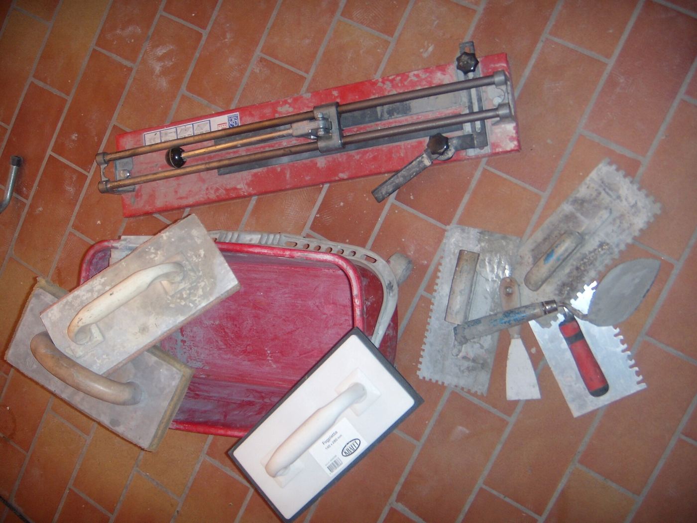Gereedschap van een tegelzetter op een rode tegelvloer: tegelsnijder, lijmkammen, spanen en een voegbord